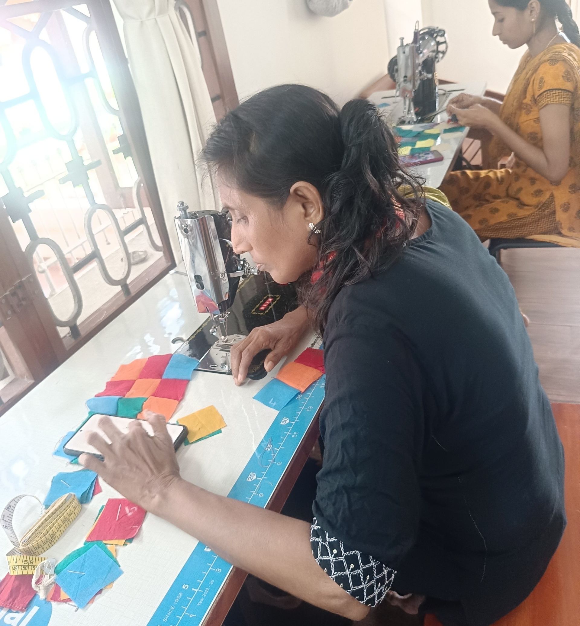 A maker lays small squares of cloth in a pattern at a sewing table. Another maker sits at a machine behind.