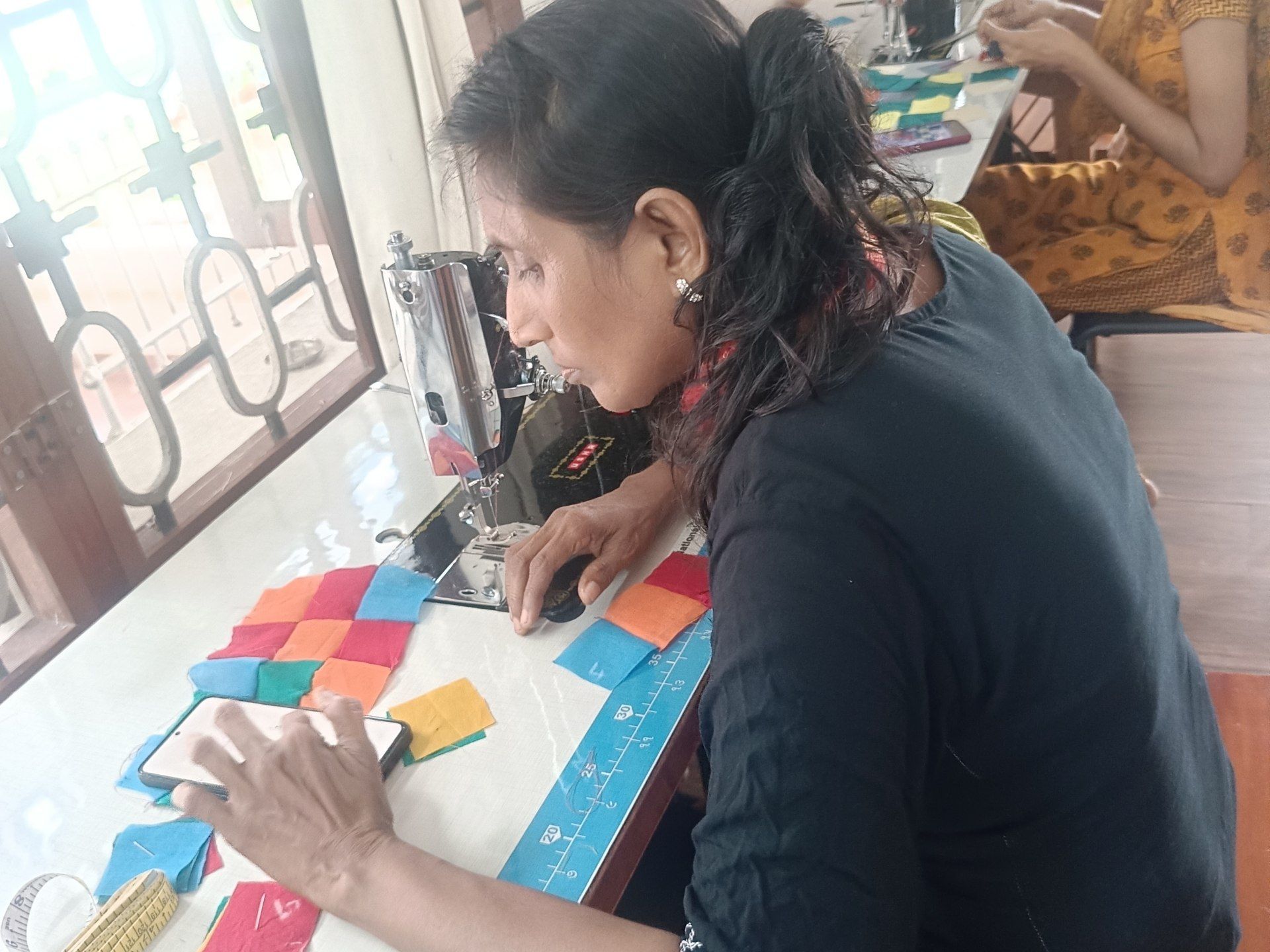 A maker lays small squares of cloth in a pattern at a sewing table. Another maker sits at a machine behind.