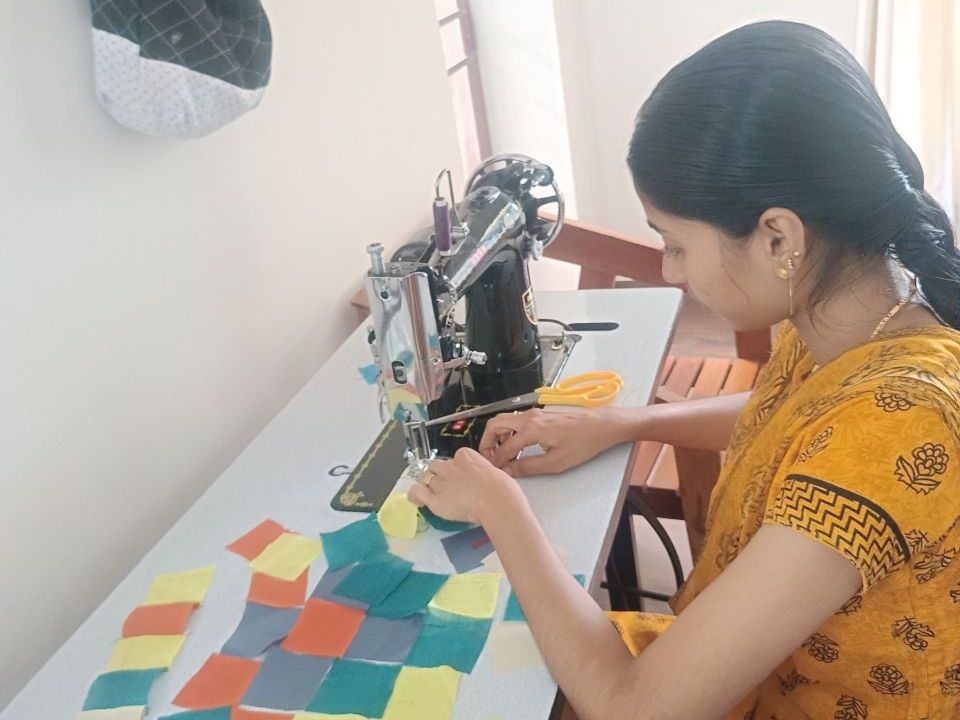 A maker works at a sewing machine. Squares of cloth lie on the table, and a phone shows a pattern.