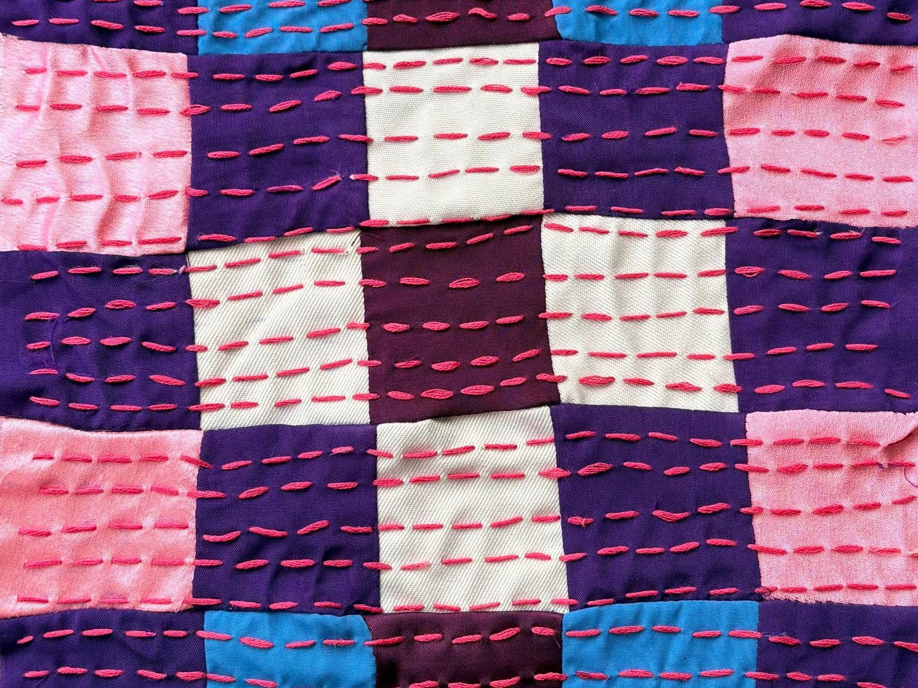Close-up of pink, purple, white and blue squares crossed by rows of red running stitch.
