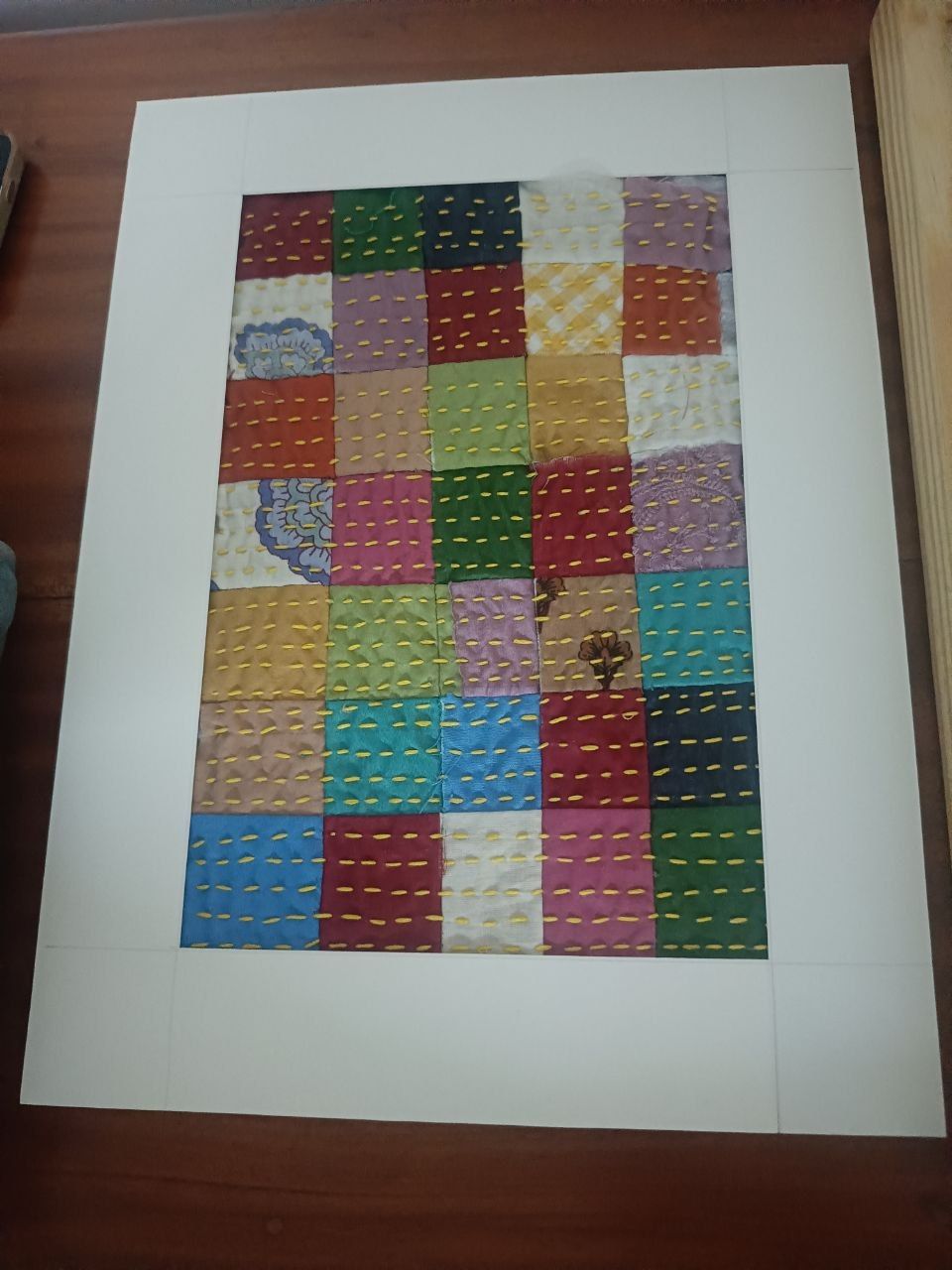 A panel of coloured squares with rows of stitches, in a white mount on a wooden table.