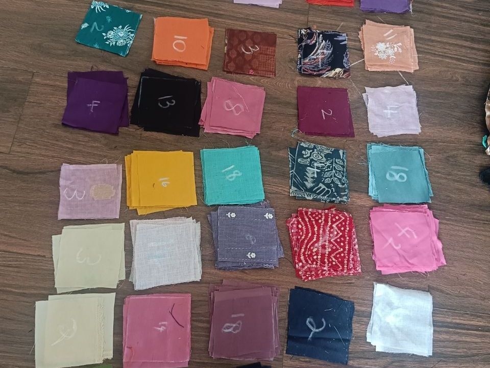 Stacks of cut cloth squares in many colours, sorted by cloth and numbered in chalk, laid in rows on a wooden floor.