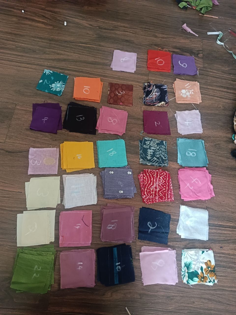 Squares of cloth in many colours laid out in rows on a wooden floor, each marked by hand.