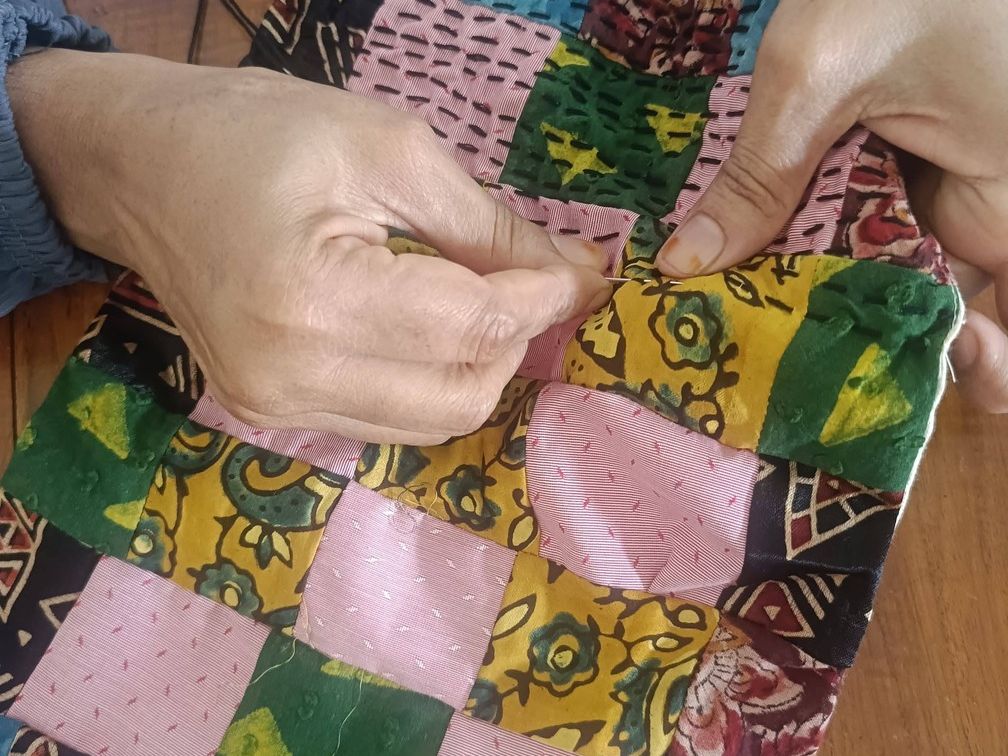 Two hands sew rows of running stitch through squares of printed and plain cloth.