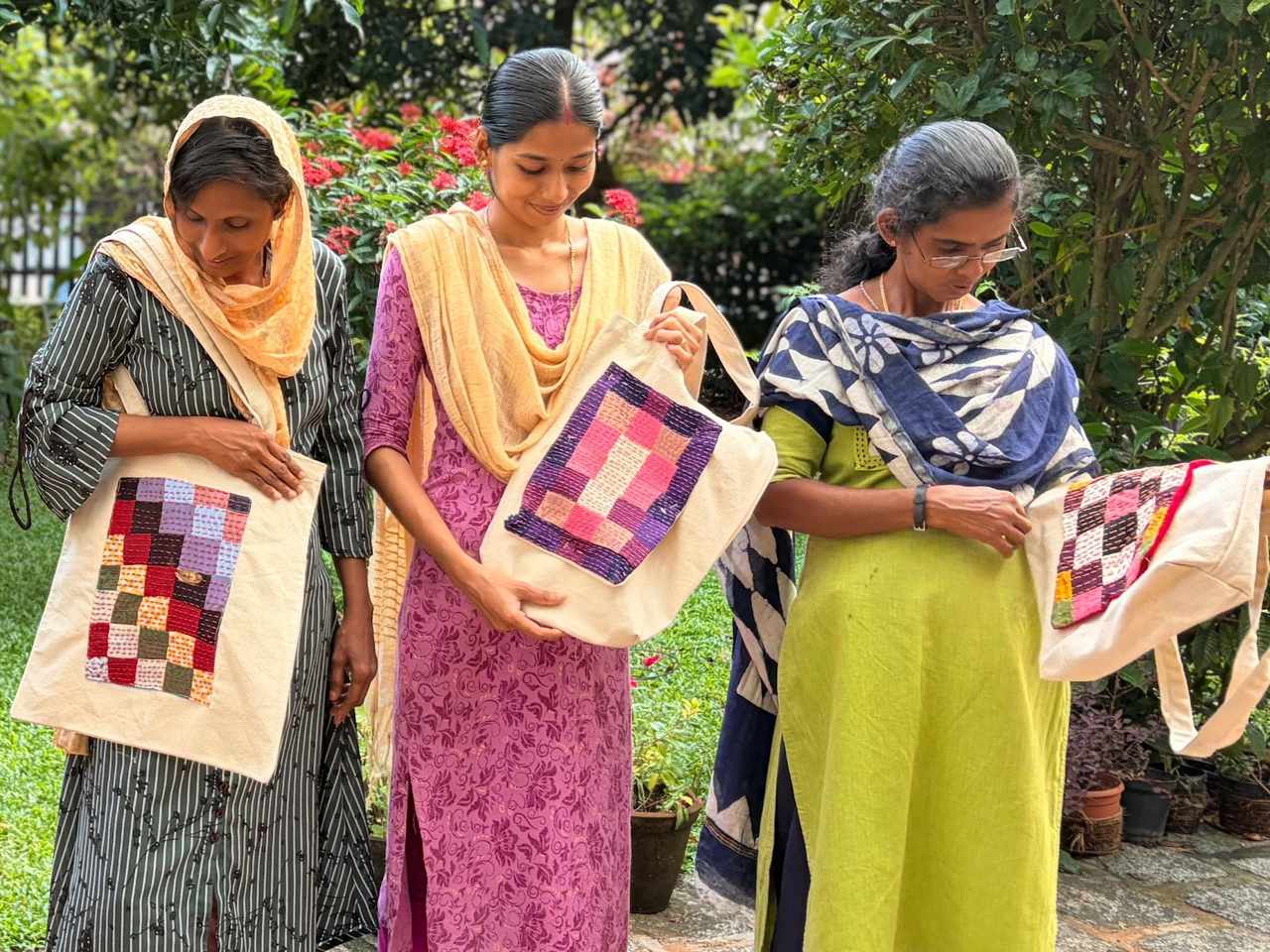 Three makers in a garden hold cotton tote bags, each with a stitched patchwork panel on the front.
