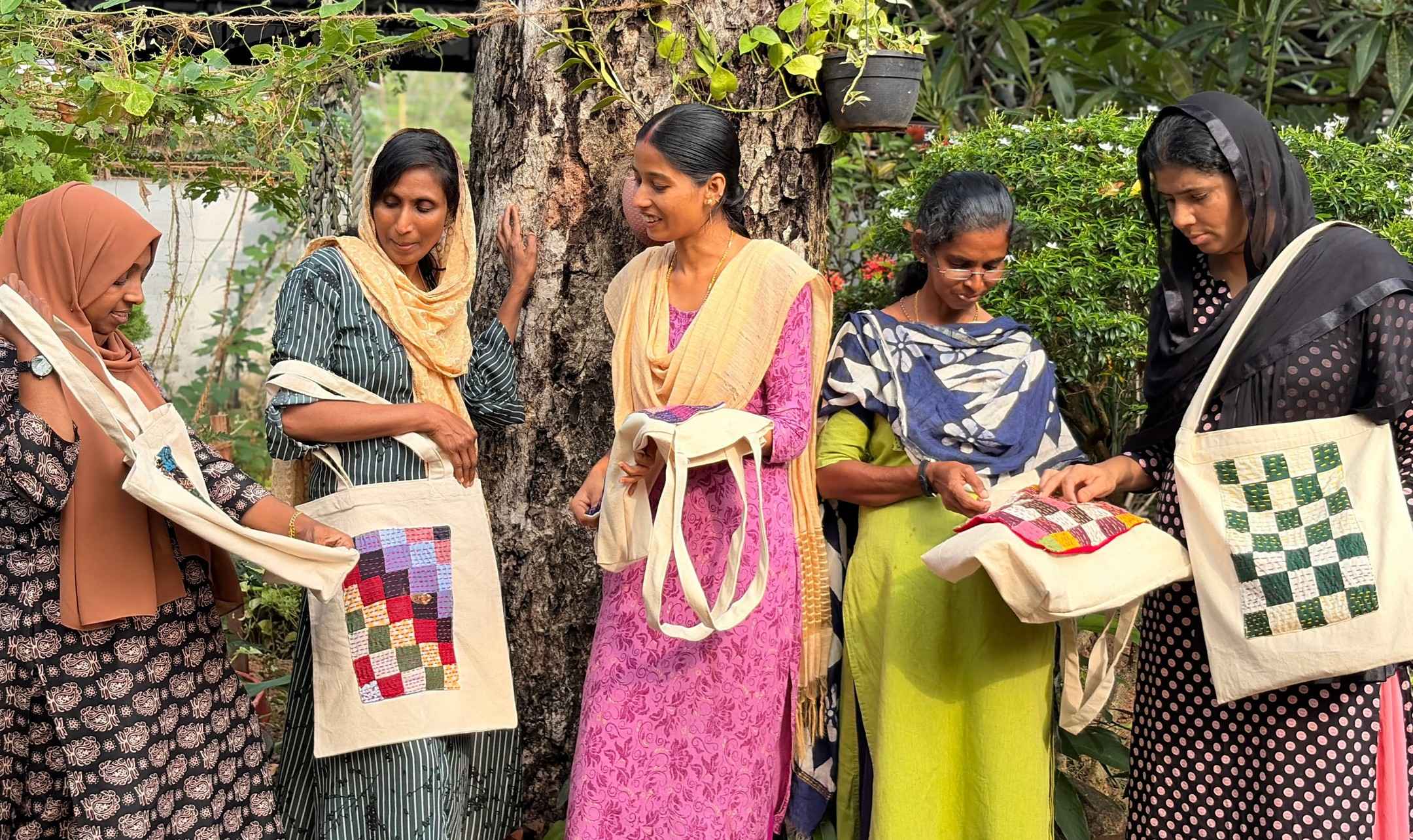Five makers stand together in a garden, each holding a cotton tote bag with a stitched patchwork panel. The panels are purple, red and green squares, a green and cream chess pattern, and a pink design.