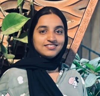 Shabana K H smiles at the camera. She wears a black headscarf and a grey-green printed top. Behind her are leafy plants and a carved wooden wall.